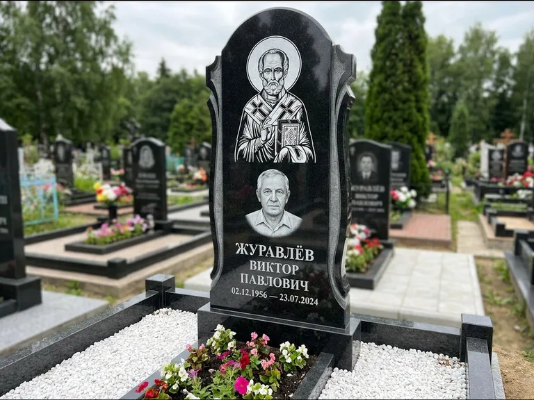 Памятник с гравировкой иконы: стела с образом в полный рост на обороте — гравировка образа Матроны Московской, низкие кусты спиреи в цвету, работа «Лебедева» в Тюмени